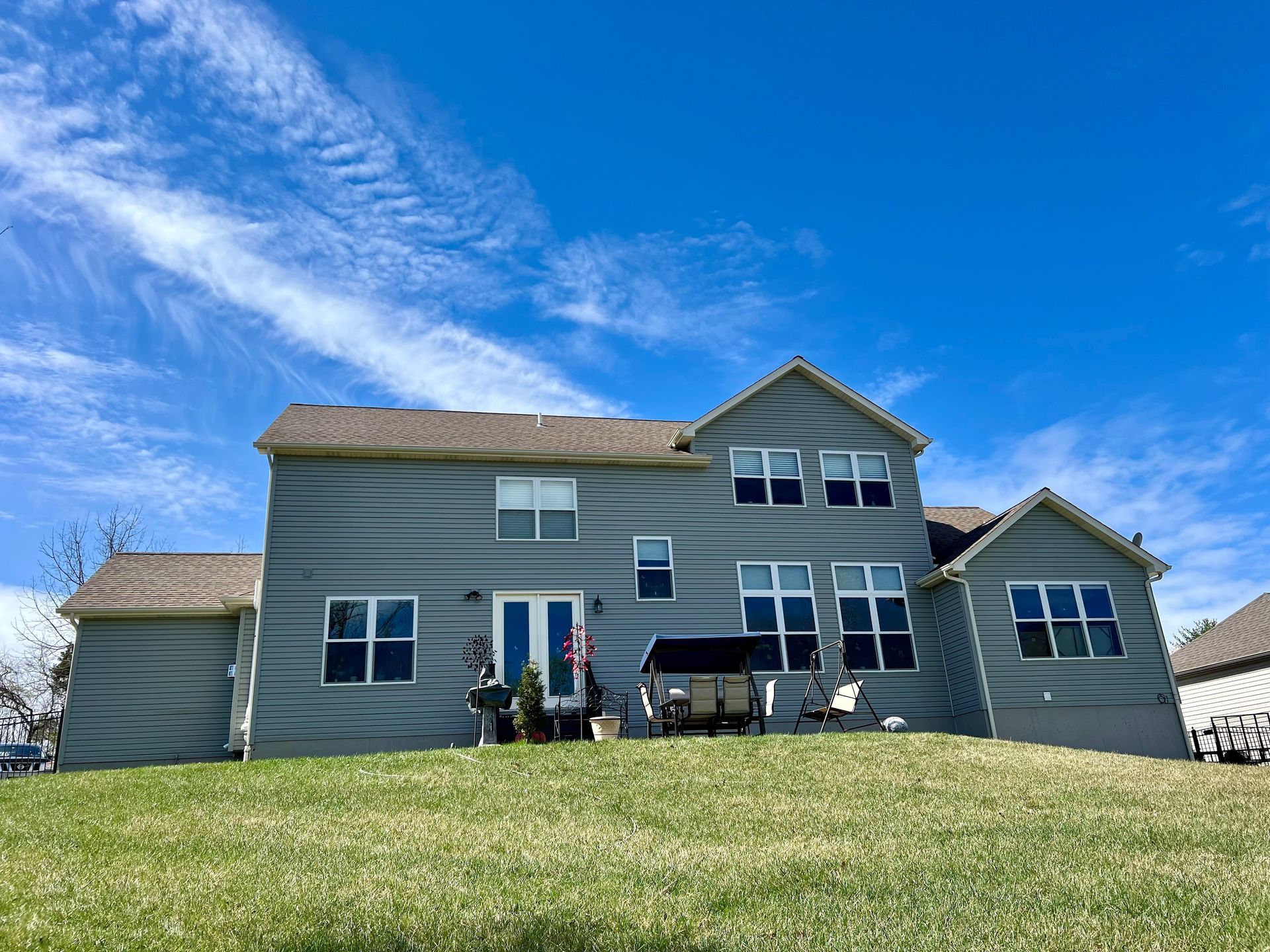 A light gray two-story house with a brown roof sits atop a grassy hill under a bright blue, cloud-streaked sky.