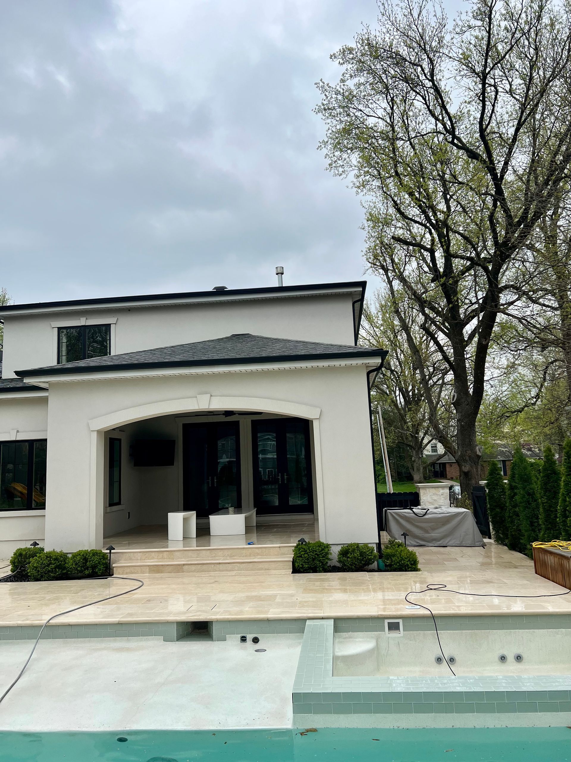 A modern two-story home with a covered patio, travertine pool deck, and an unfinished pool under a cloudy sky.