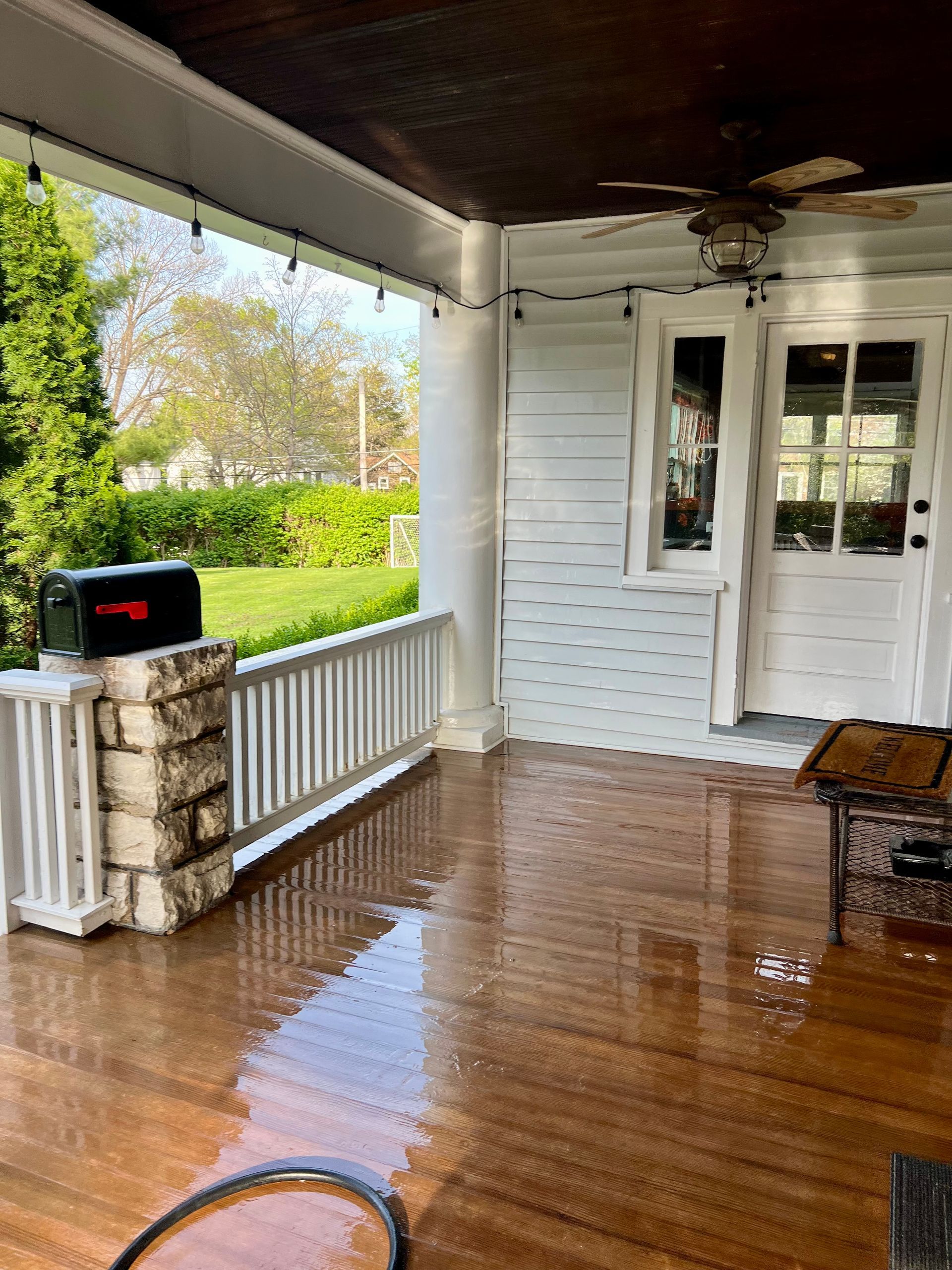 A wet wooden porch with white railings, a stone pillar with a black mailbox, and a white door with glass panels.