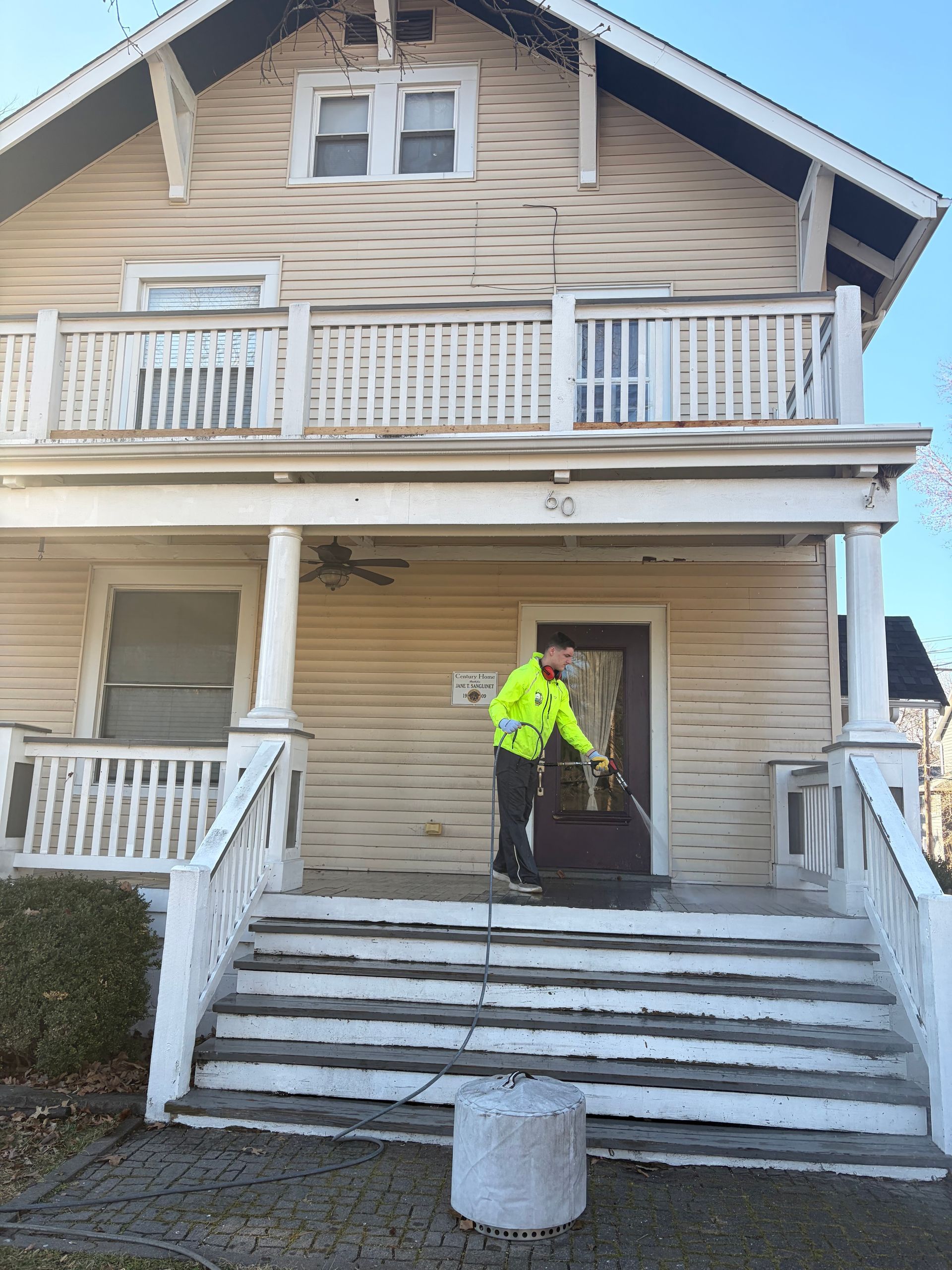 A person in a bright yellow jacket stands on the front porch steps of a light-colored, two-story house.