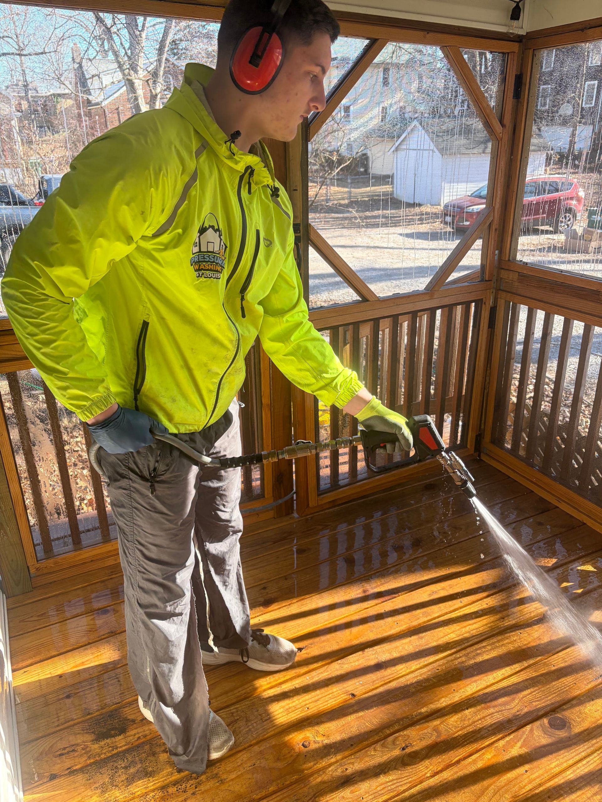 A person in a neon yellow jacket and ear protection pressure washes a wooden deck inside a screened-in porch.