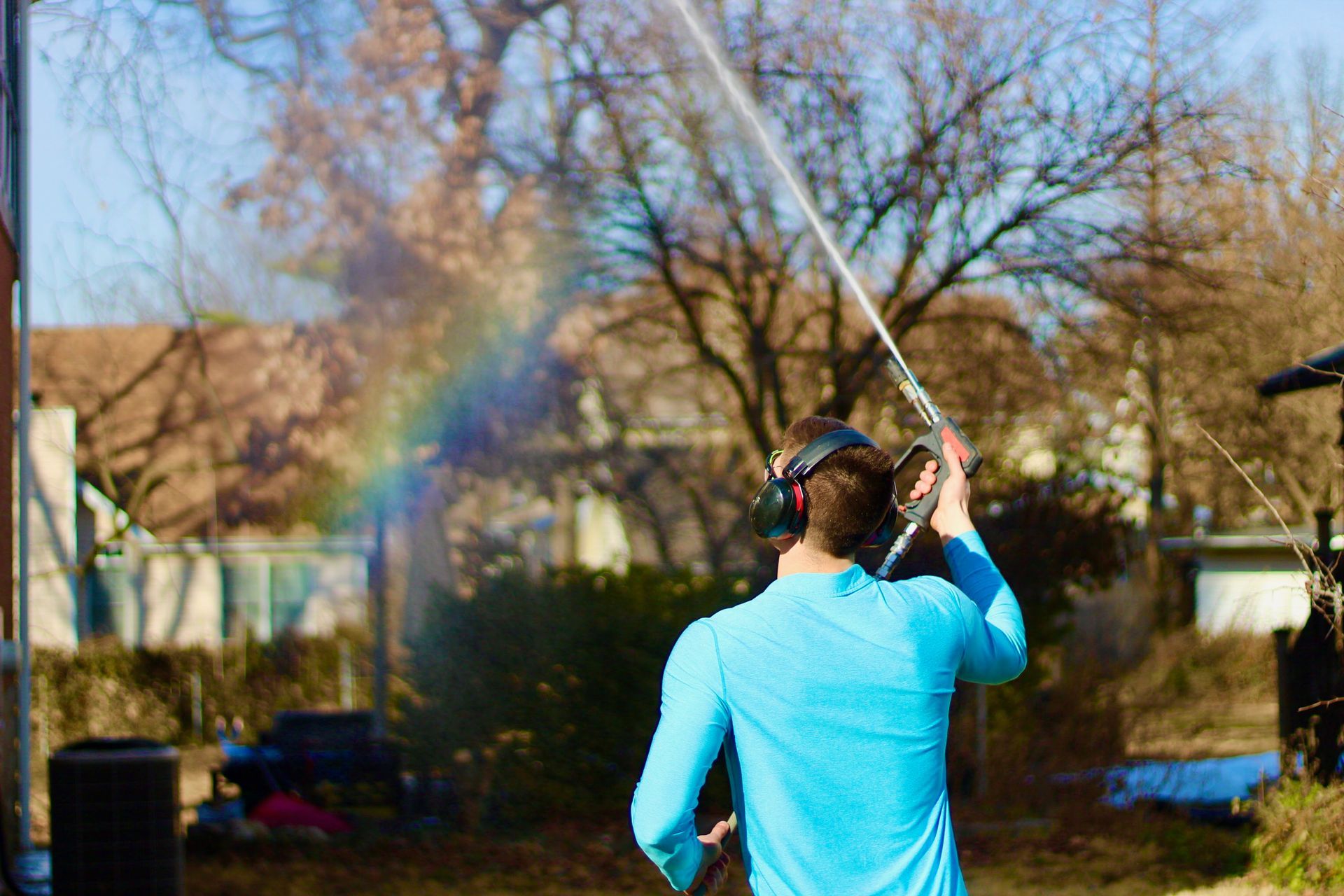 A person in a bright blue shirt wearing headphones uses a pressure washer, creating a rainbow in the water spray.