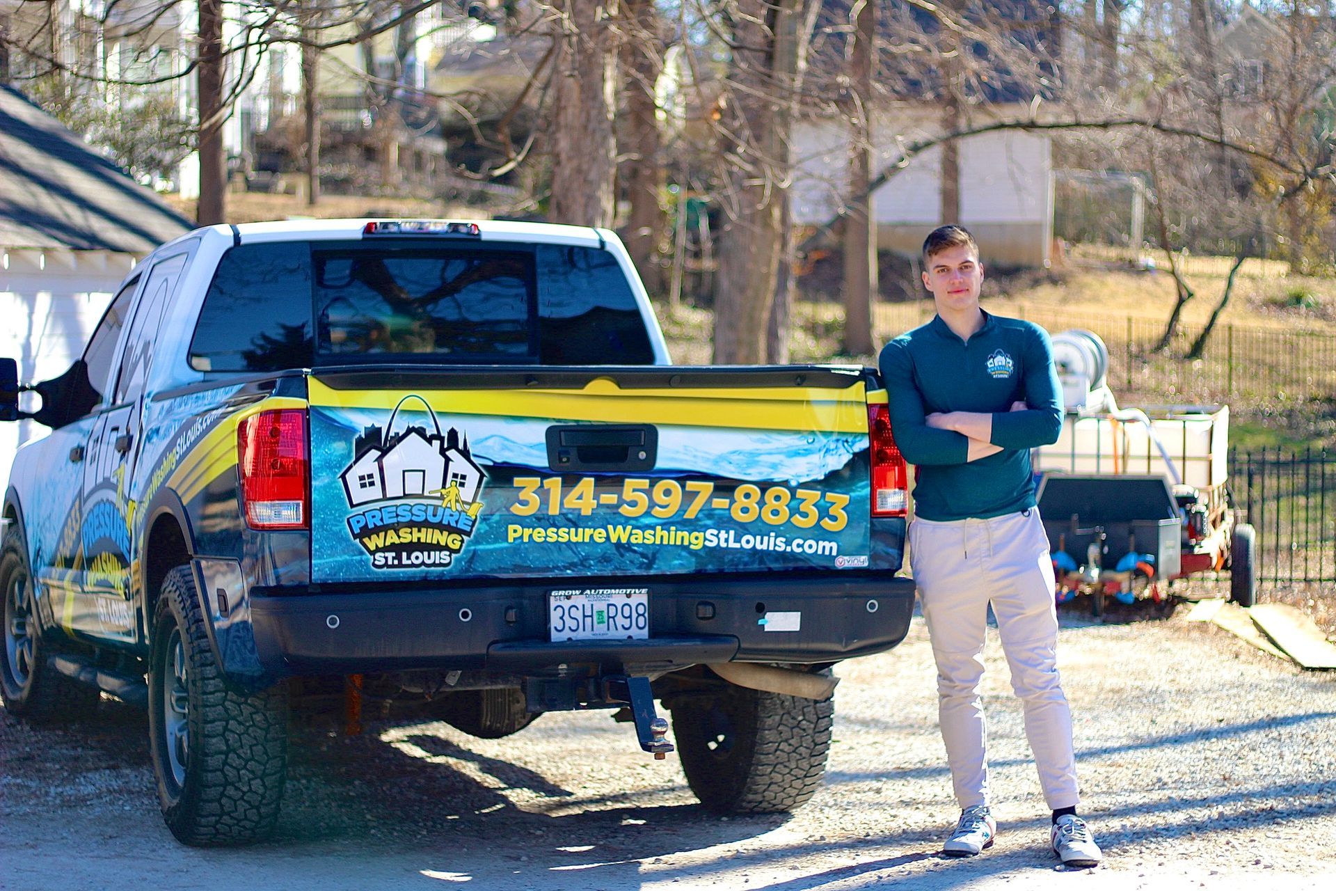 A person stands with arms crossed next to a pickup truck featuring a branded advertisement for a pressure washing service.