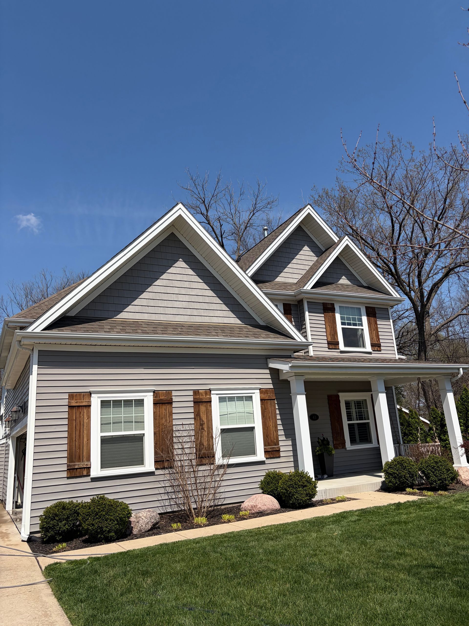 A gray two-story house with a peaked roof, white trim, and brown shutters, set against a clear blue sky.