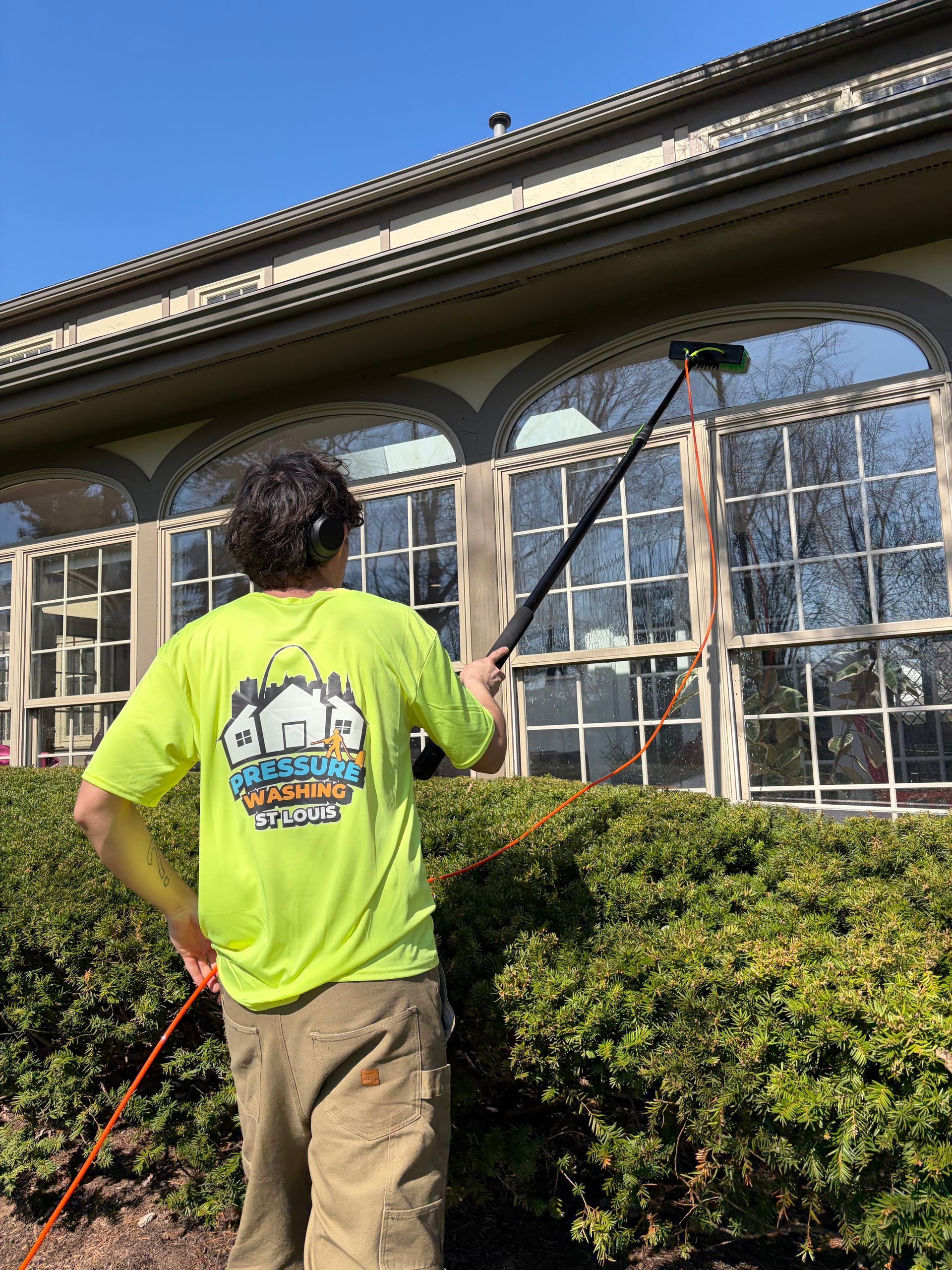 A person in a neon yellow shirt cleans large, arched windows on a building exterior using a long-handled brush.