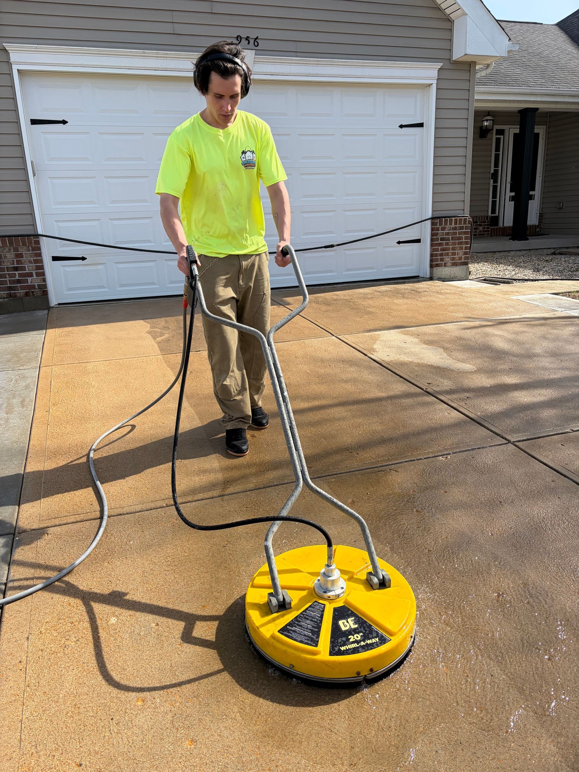 A worker in a bright yellow shirt uses a yellow rotary surface cleaner to pressure wash a concrete driveway.