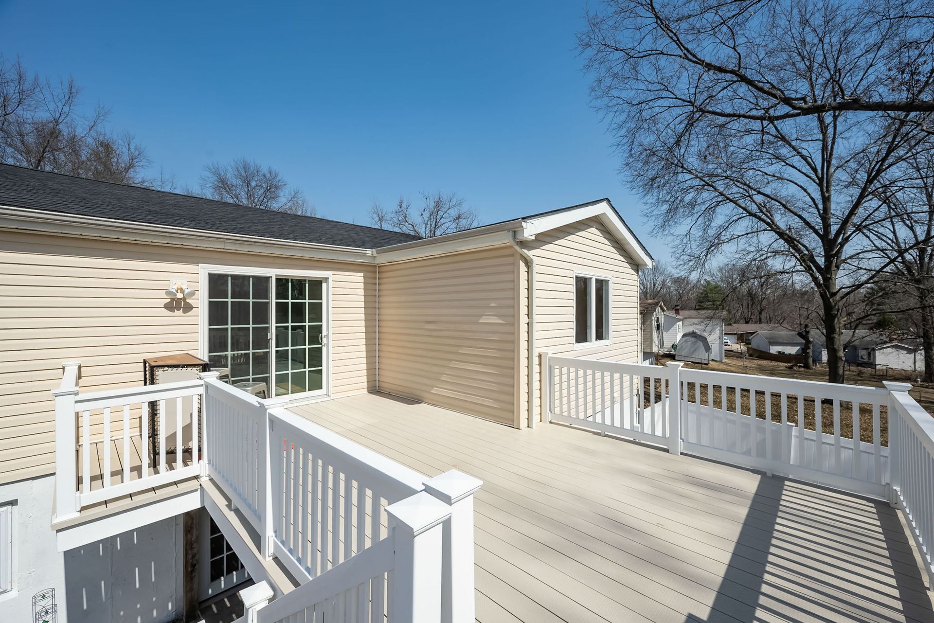 A beige house with a spacious wooden deck, white railings, and a sliding glass door under a clear blue sky.