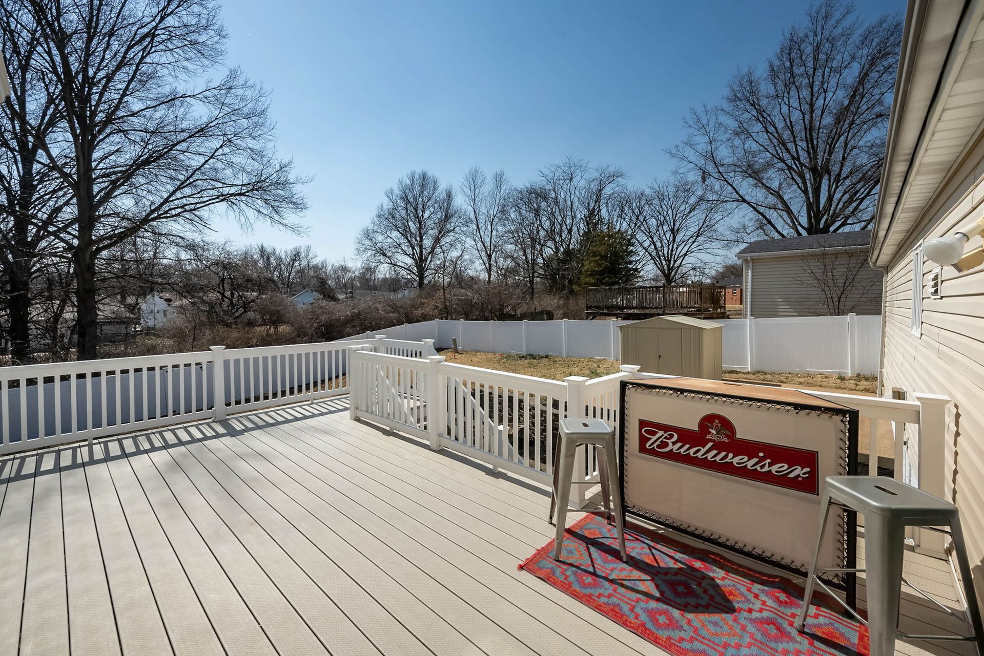 A sunlit wooden deck featuring a Budweiser-branded bar, two metal stools, and a patterned rug, set against a fenced yard.