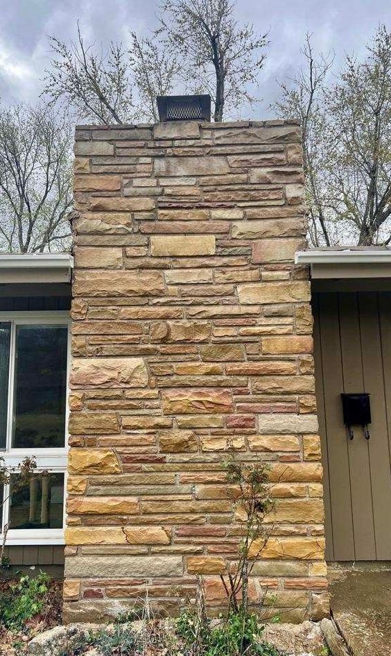 A exterior view of a tall, rectangular chimney built from horizontal tan and brown stacked fieldstone.