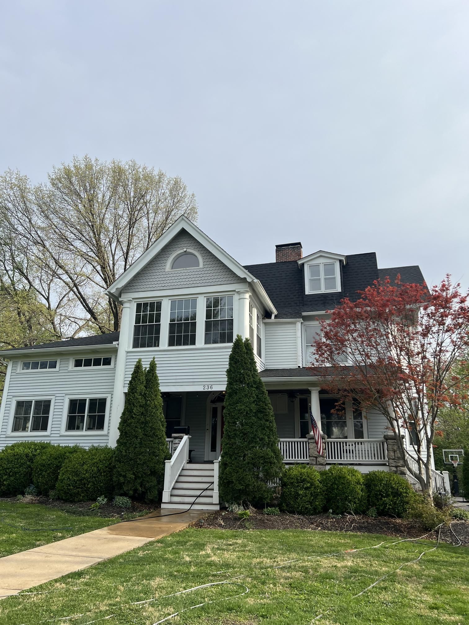 A two-story white house with a gable roof, front porch, and tall green hedges under a cloudy sky.