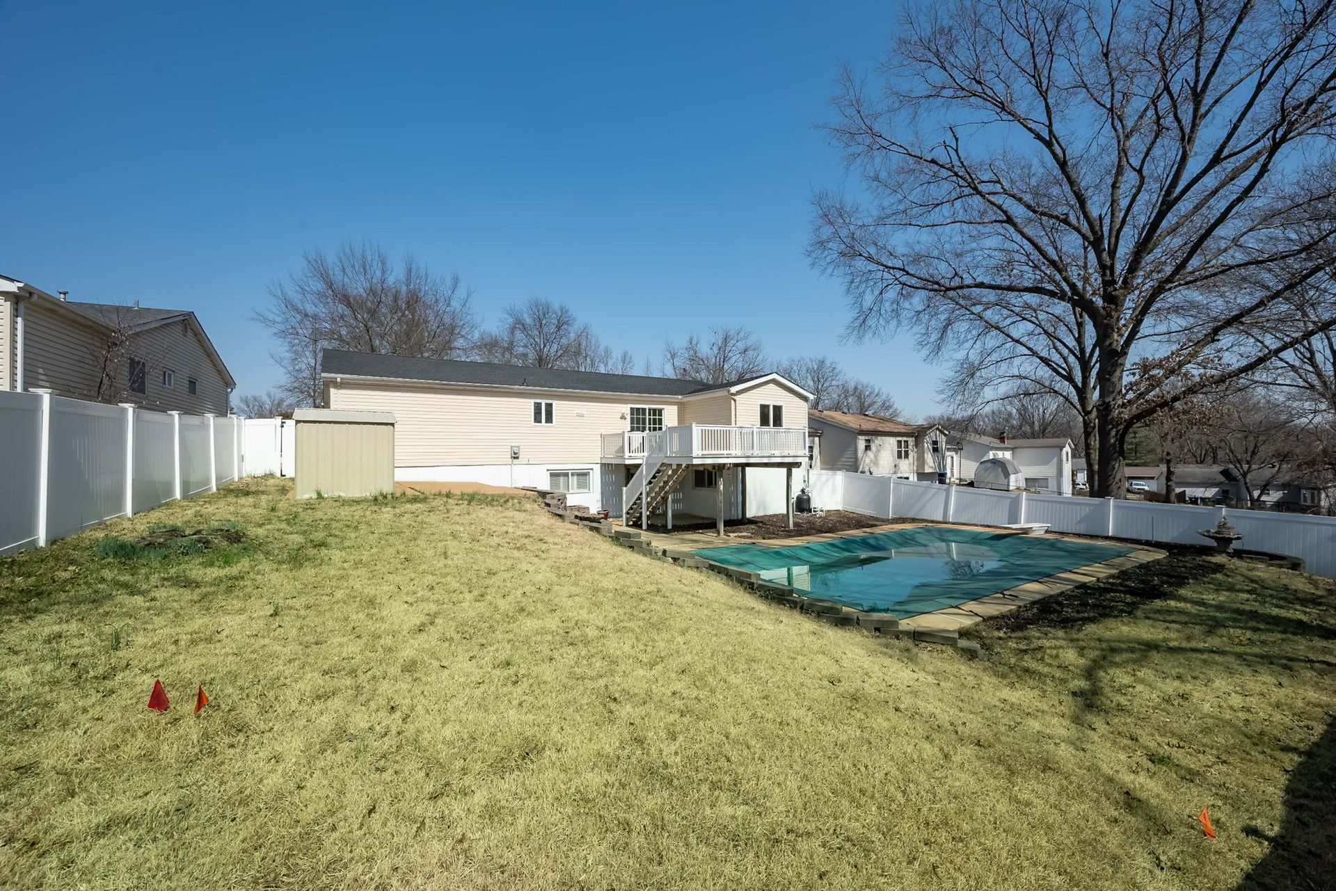 A backyard with a green lawn, a covered swimming pool, a small shed, and a two-story house with a white deck.