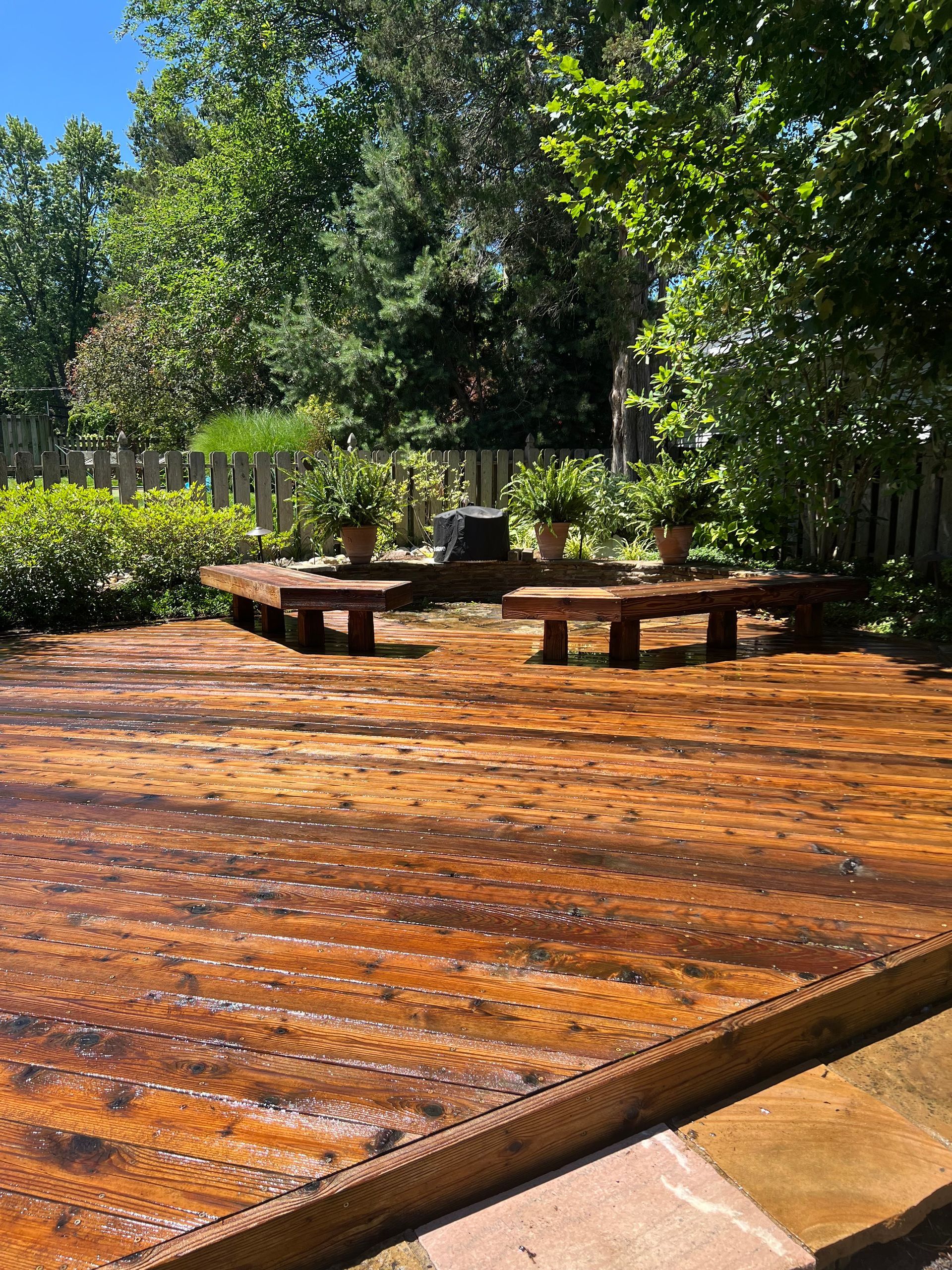 A stained wooden deck with a built-in semi-circular bench, set in a sunny, tree-filled backyard.