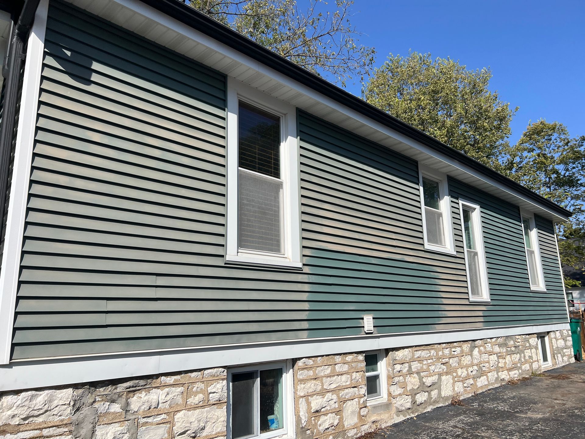A side view of a house with light green horizontal siding and a stone foundation featuring three basement windows.
