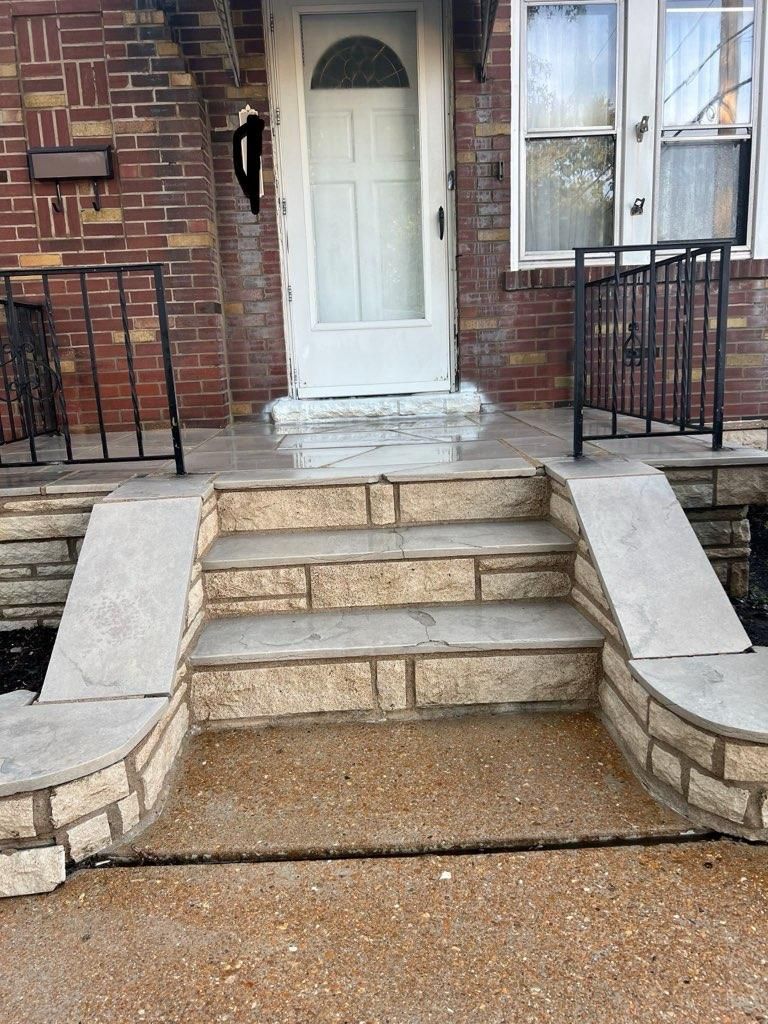 A white door sits at the top of stone steps leading to a brick house with black metal railings.