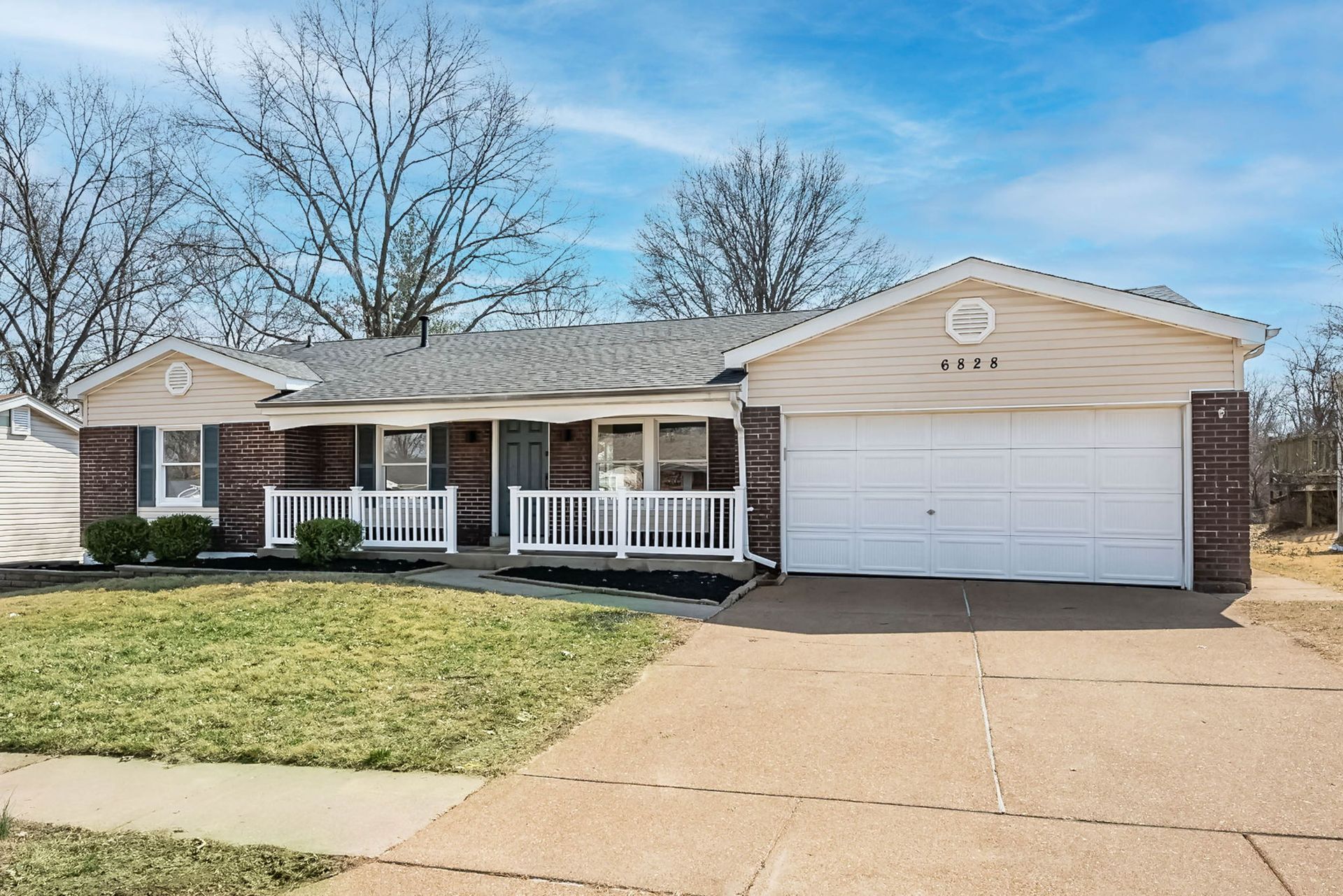 A single-story, light-colored suburban house with a brick exterior, a front porch, and an attached two-car garage.