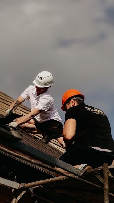 Two workers in hard hats repair a roof with shingles under a cloudy sky.