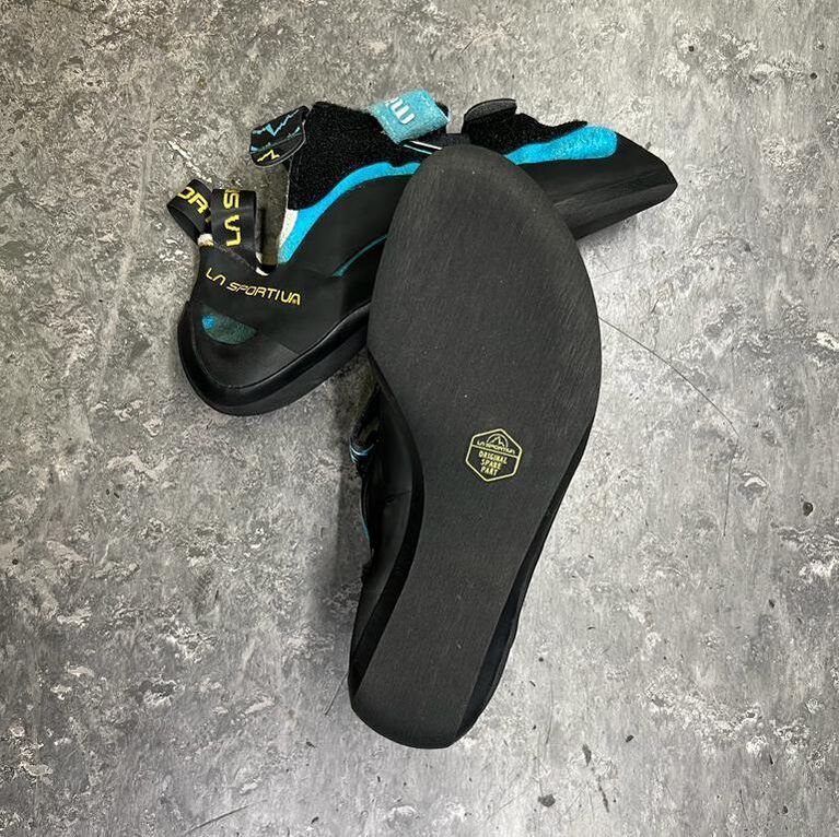 Black and teal climbing shoes, one sole facing up, on a gray speckled surface.