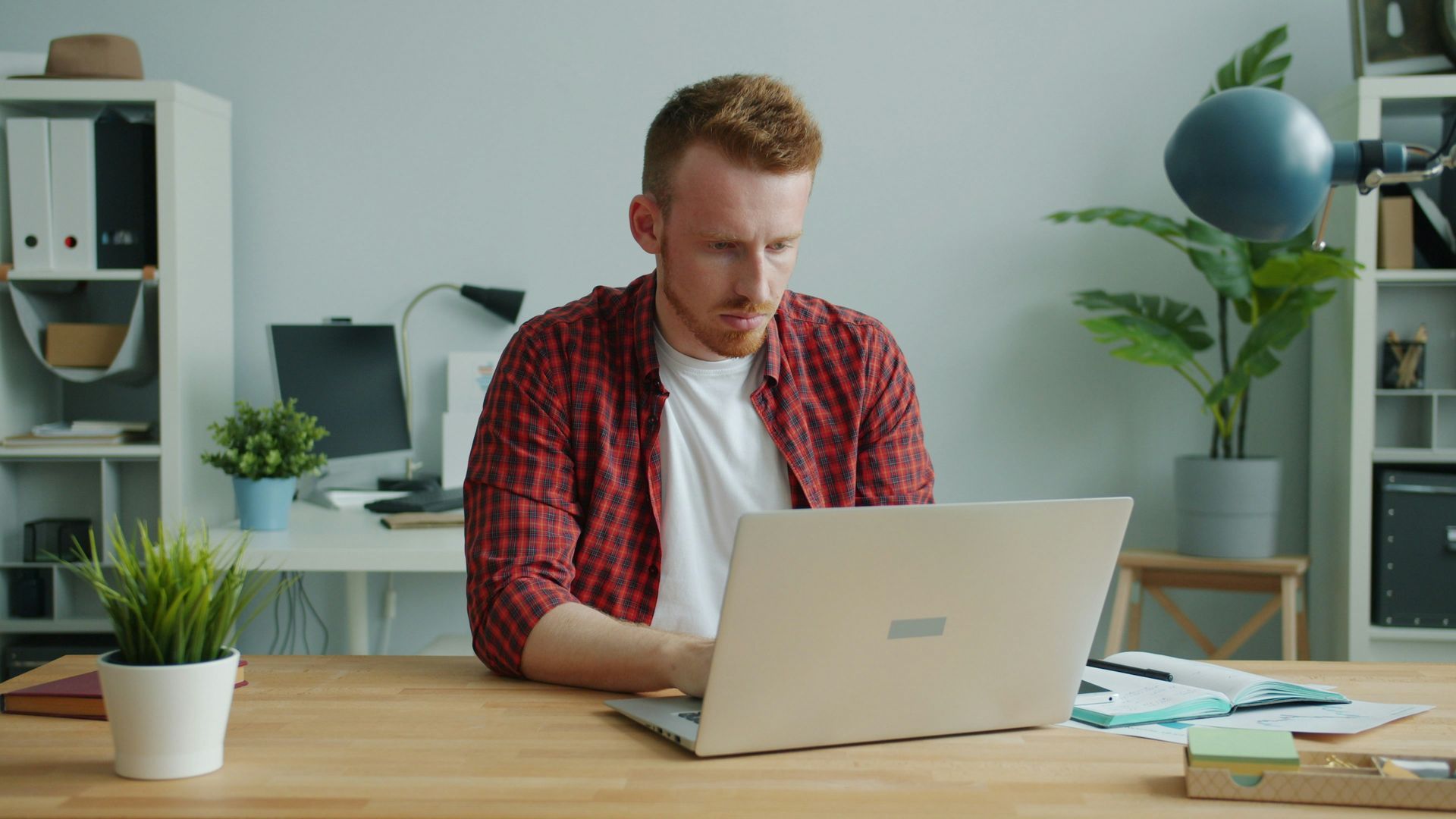Man with red hair, working on laptop at a desk in a well-lit office.