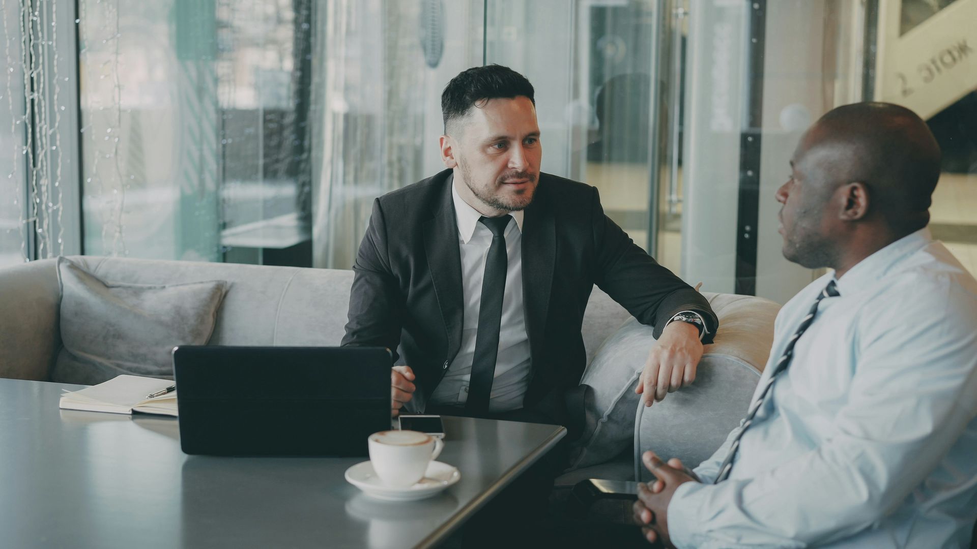 Two men in suits at a table in a modern setting, one speaking, laptop and coffee present.