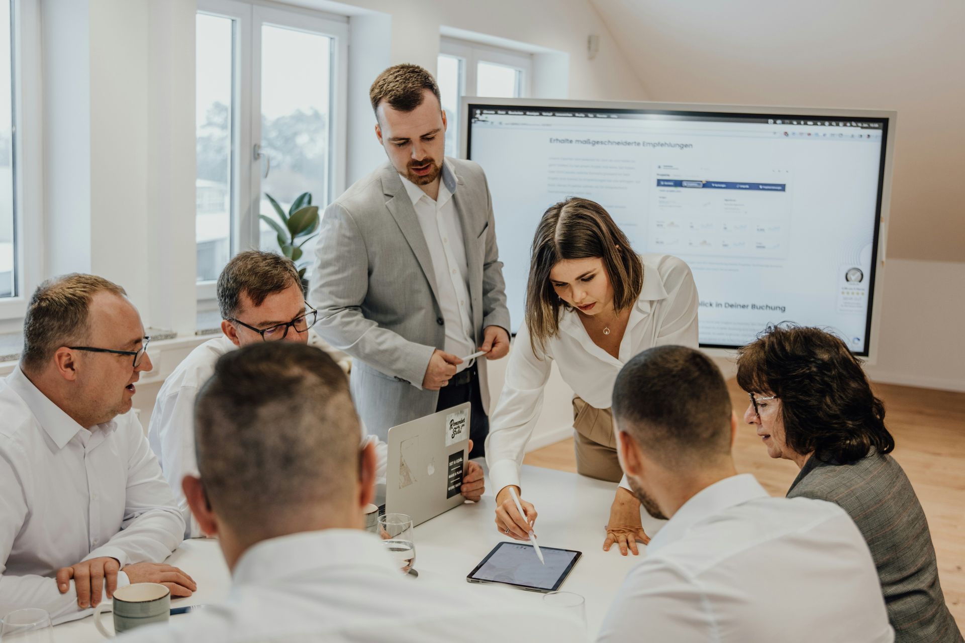 Group of professionals in an office meeting around a table, looking at a tablet. Whiteboard in background.