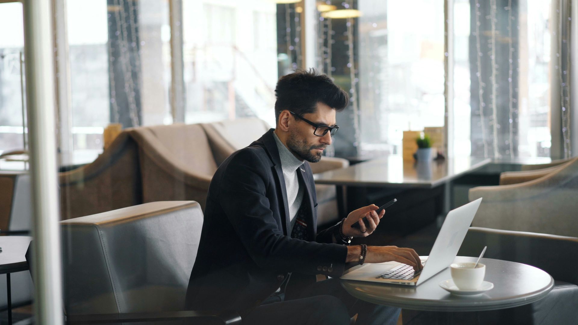 Man in suit working on laptop, holding phone at a table in a cafe.