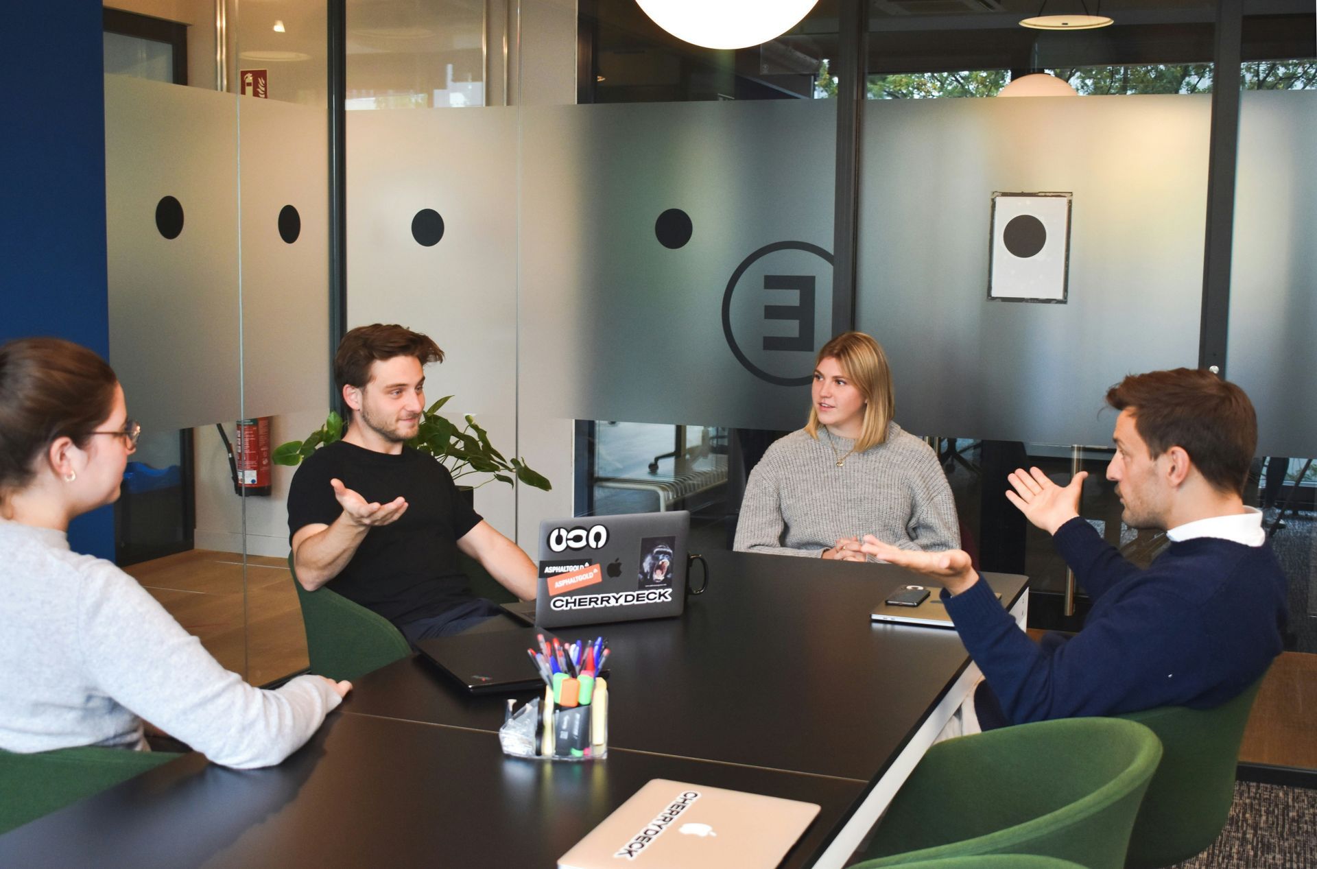 Four people in a modern office, gesturing and talking around a table. Frosted glass windows.