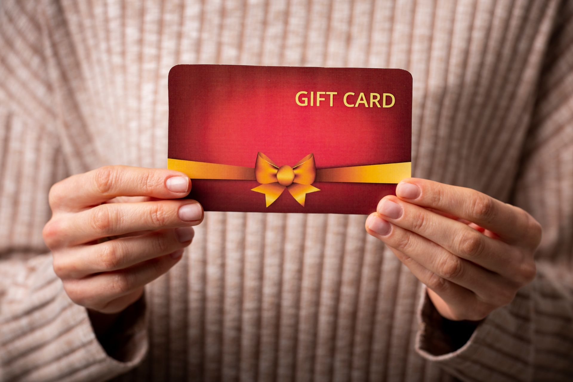 Hands holding a red gift card featuring a gold ribbon and bow against a textured beige knit sweater background.