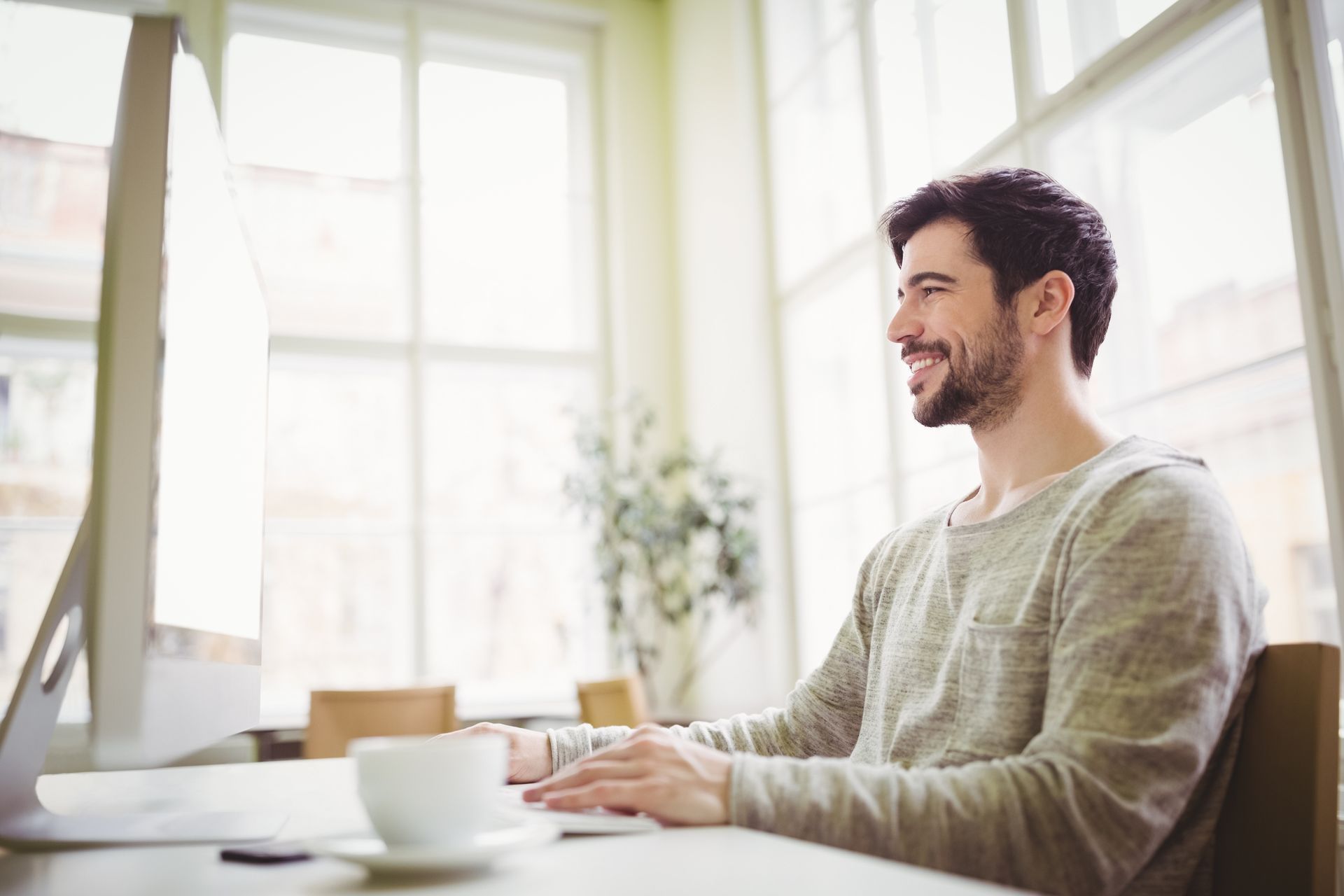 A smiling person wearing a grey sweater types on a computer in a brightly lit office with large windows.