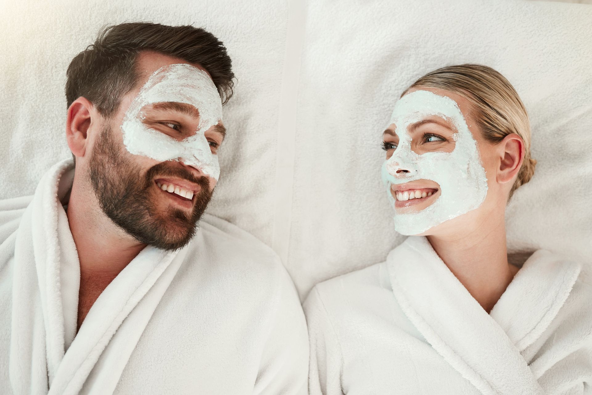 A couple wearing white bathrobes and facial masks lying on white towels, smiling at each other.
