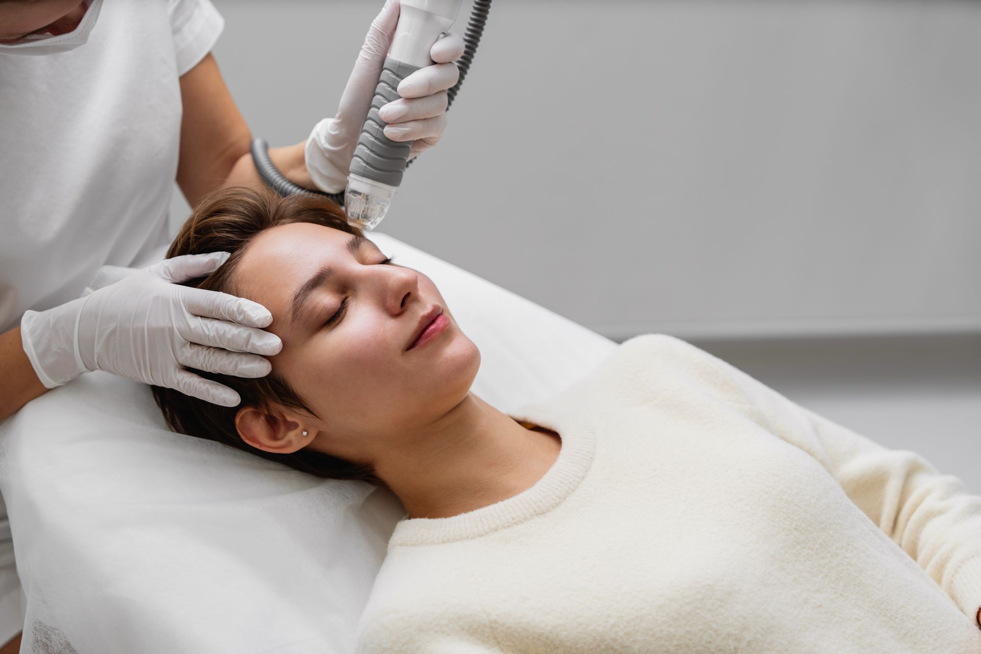 A practitioner in white gloves uses a laser device on the forehead of a patient lying on a treatment table.
