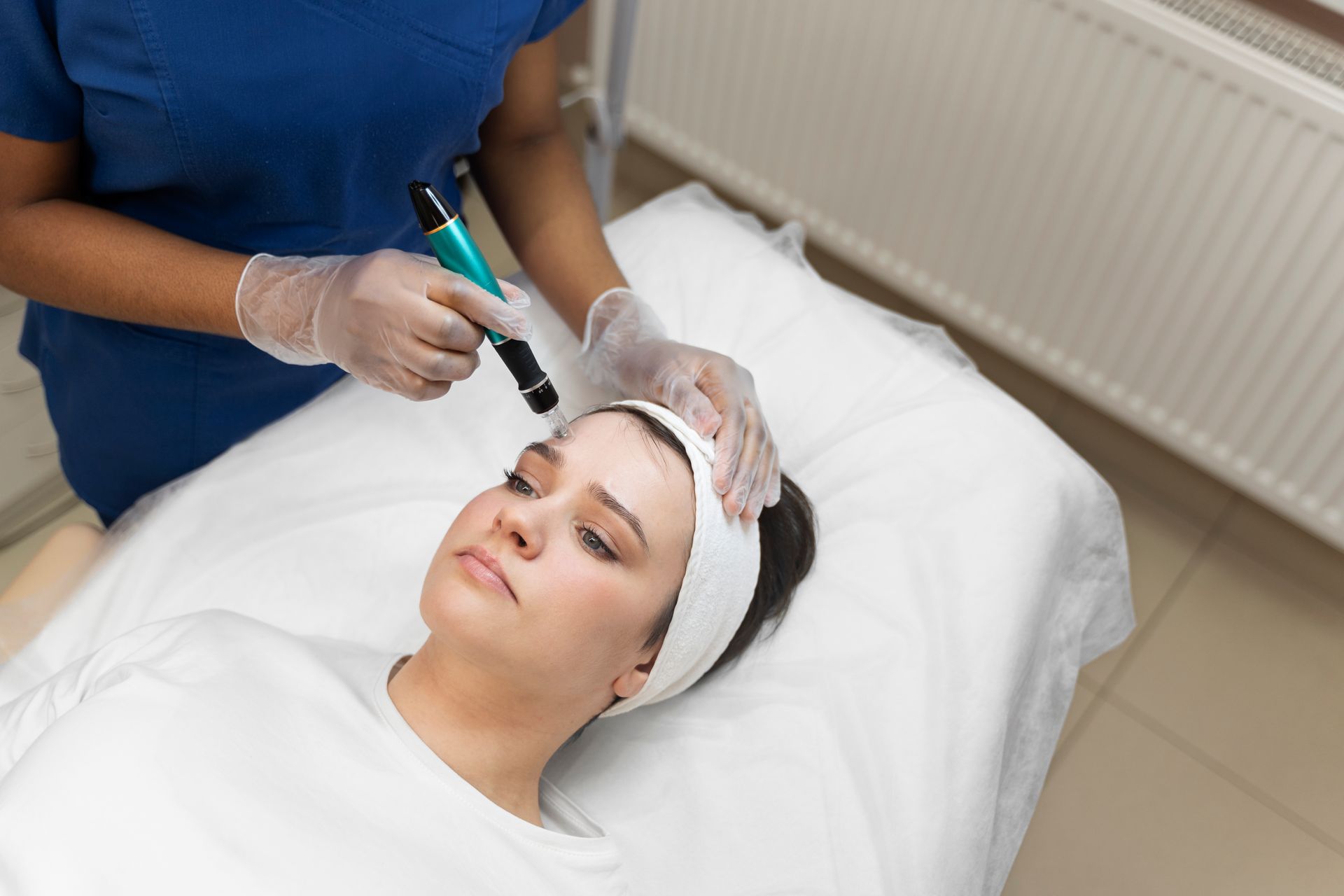 A person in medical scrubs performs a microneedling treatment on a client lying on a treatment table.