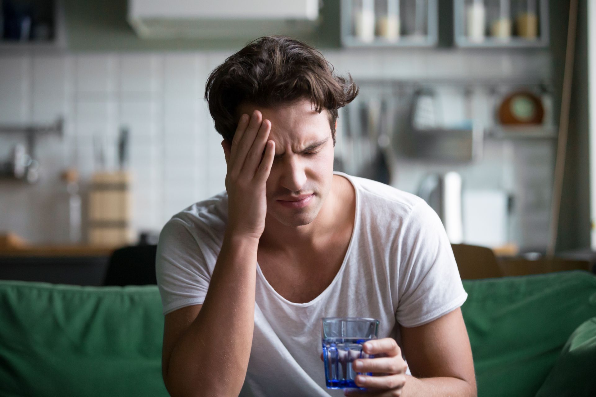 Man holding head, grimacing with a glass of water, sitting on a green couch indoors.