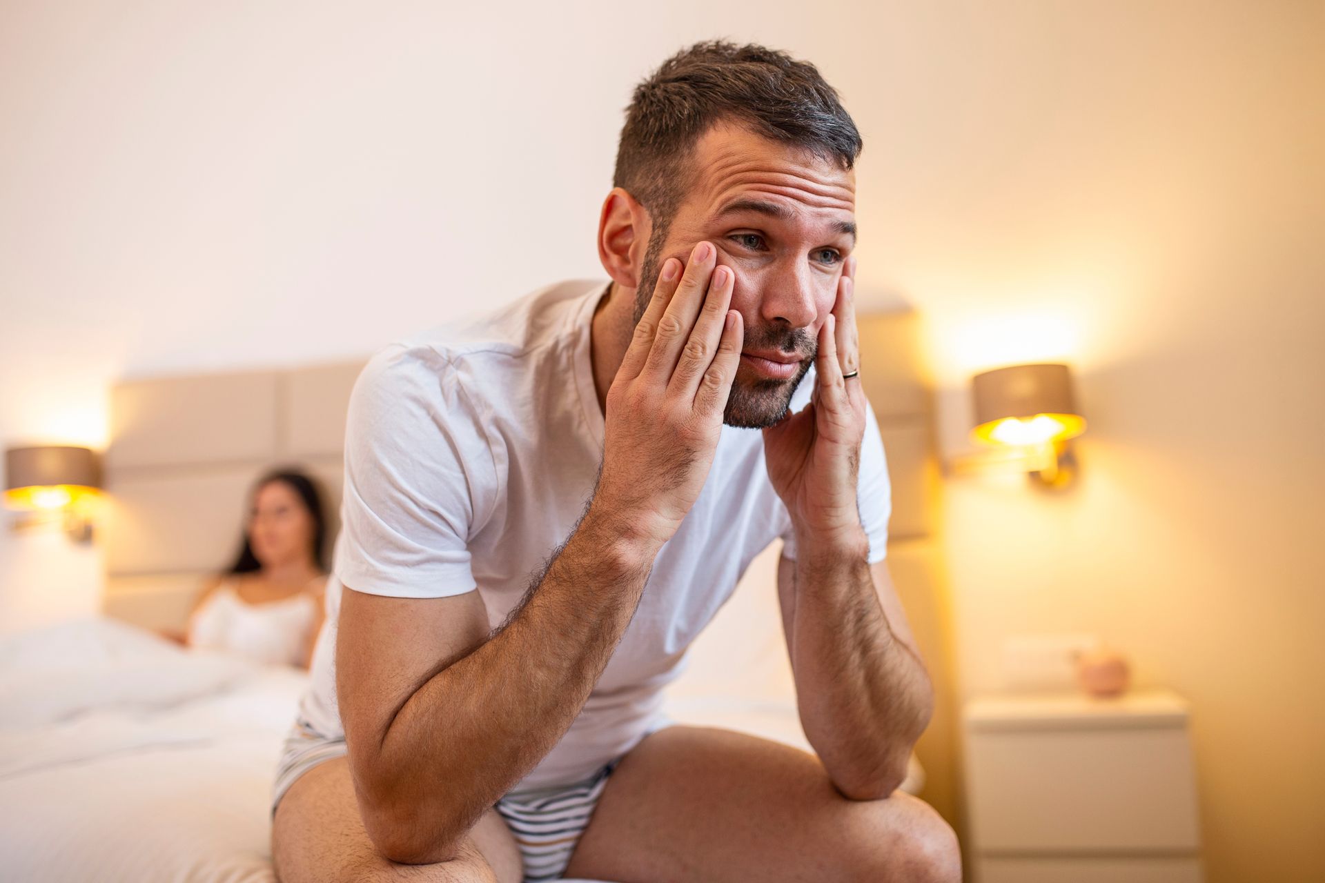 Man with worried expression sitting on a bed, woman in the background. Bedroom setting.