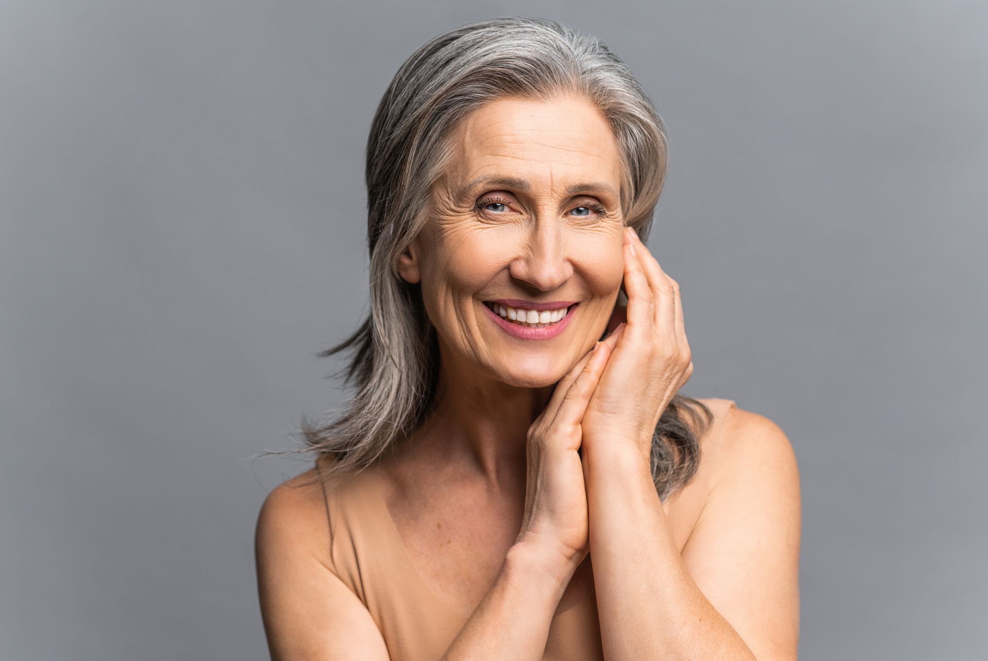 Smiling gray-haired woman posing with hands on face against a gray background