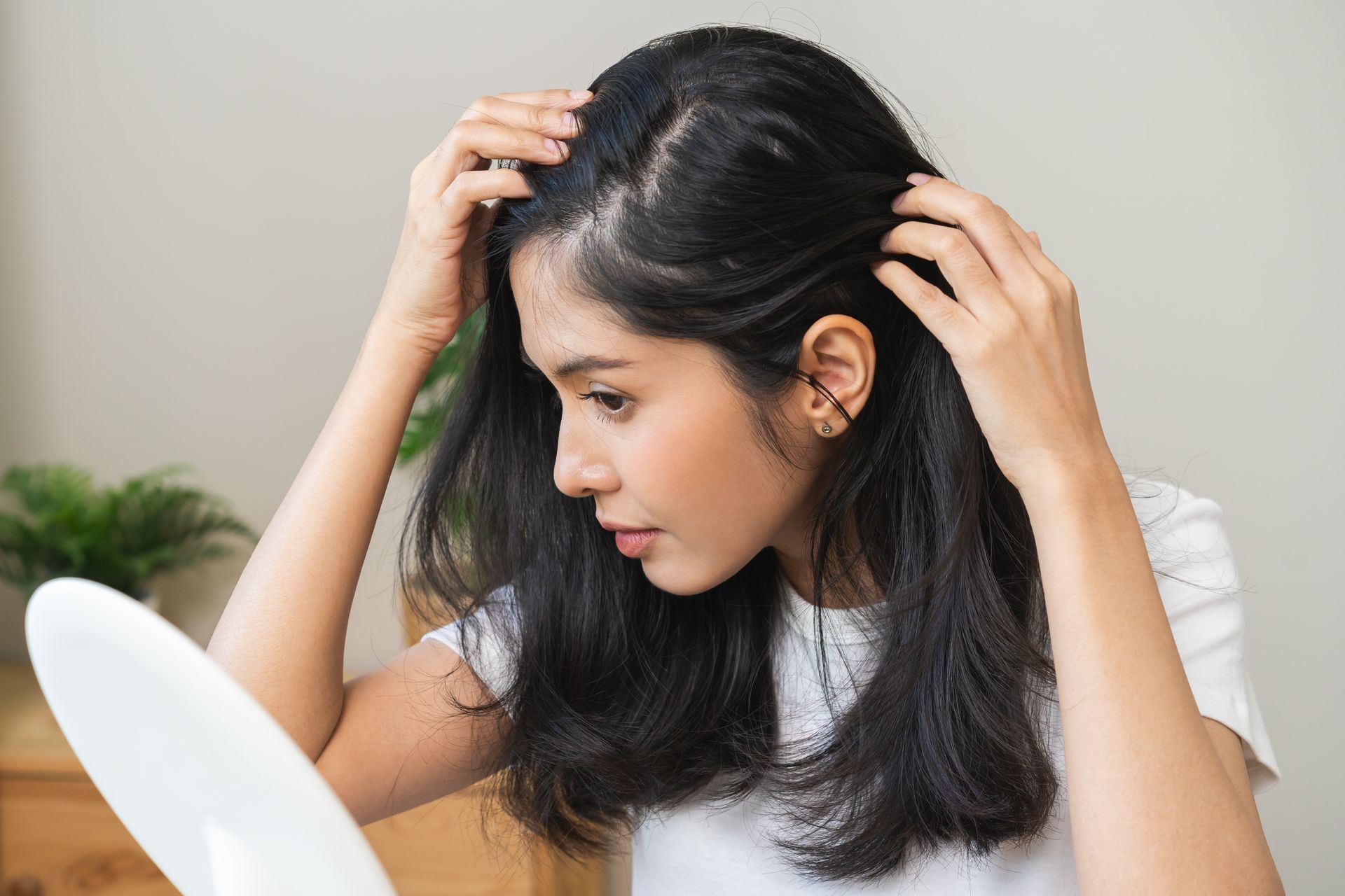 Woman examining hair for thinning in a well-lit room, holding a mirror, concerned expression.