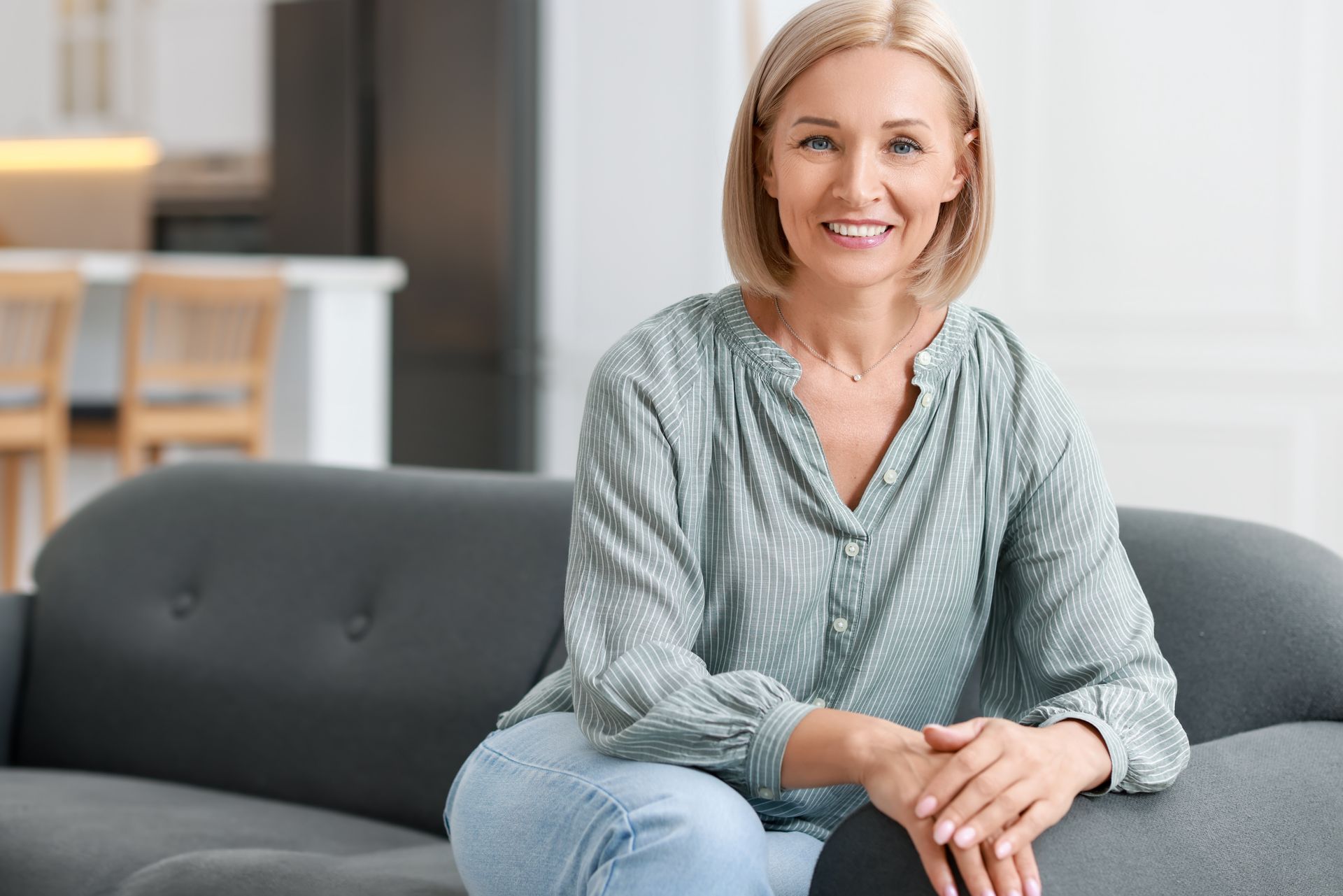 Woman with blonde bob sits smiling on a gray couch in a bright living room.