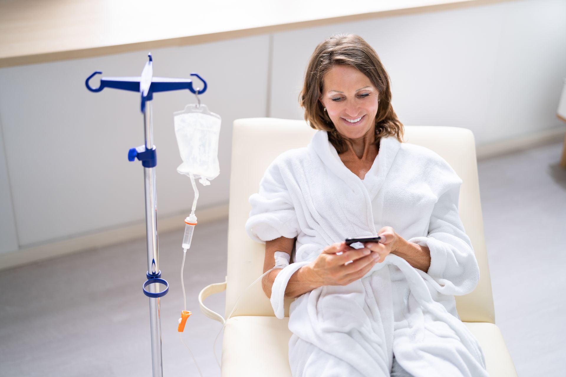 A smiling person in a white robe receives an IV drip while relaxing in a chair and using a smartphone.