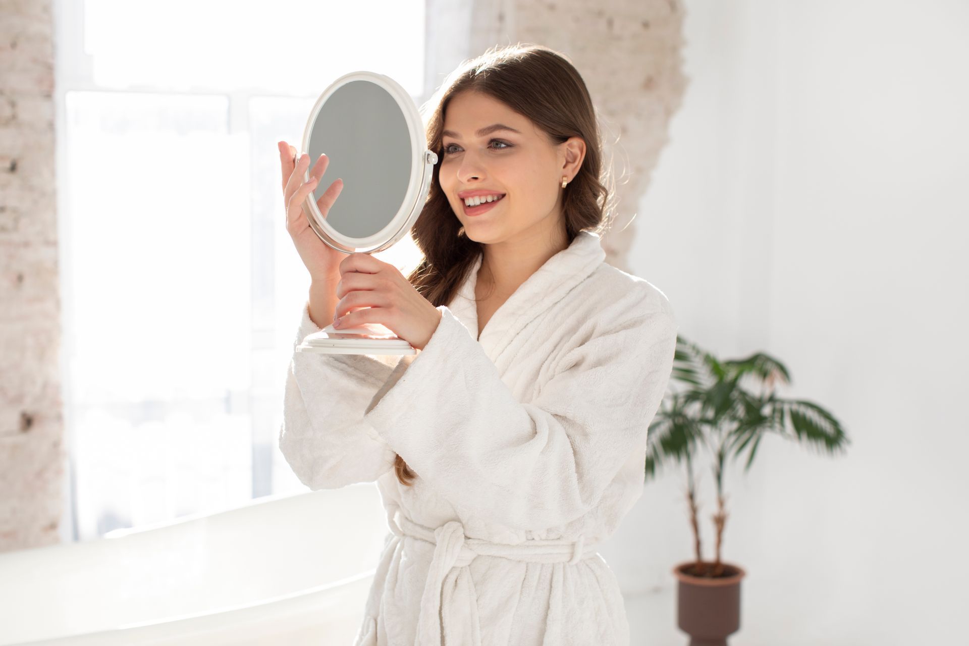 Woman in white robe smiles, looking in a handheld mirror in a bright, white room with a plant.