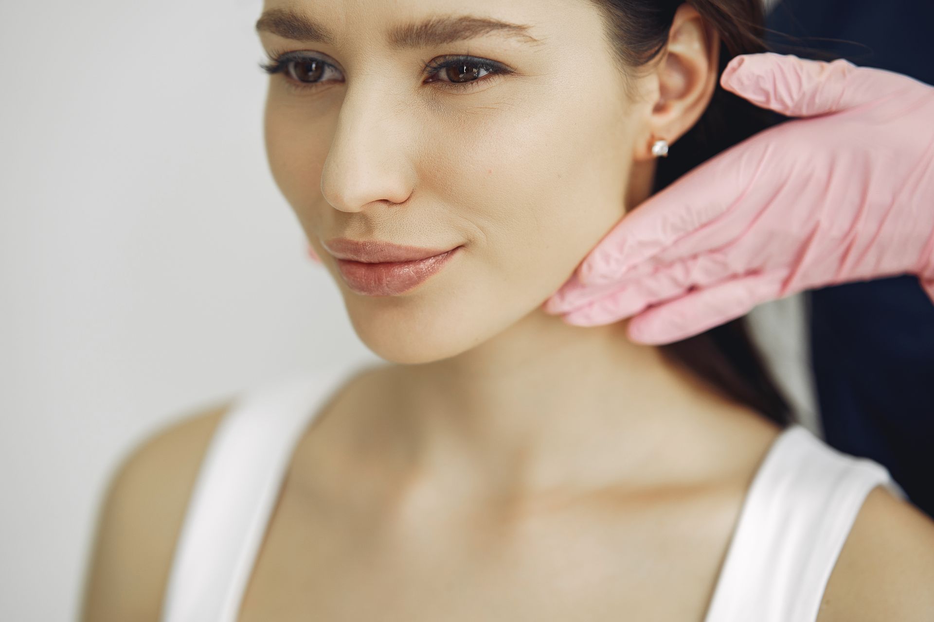 A person in a white tank top receives a gentle examination of their jawline from a hand wearing a pink medical glove.