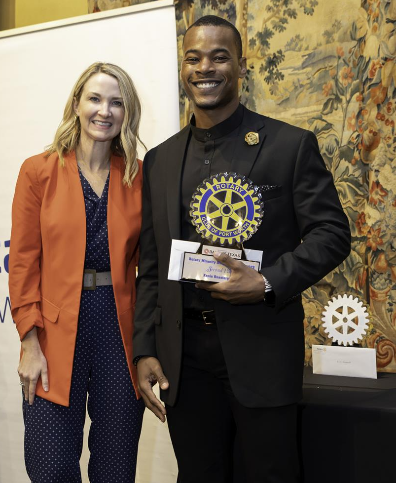 Man in black suit holding award, stands next to woman in orange blazer and blue jumpsuit, Rotary Club event.