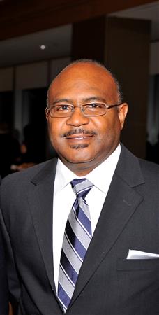 Man in a dark suit and striped tie smiles, wearing glasses in a formal setting.