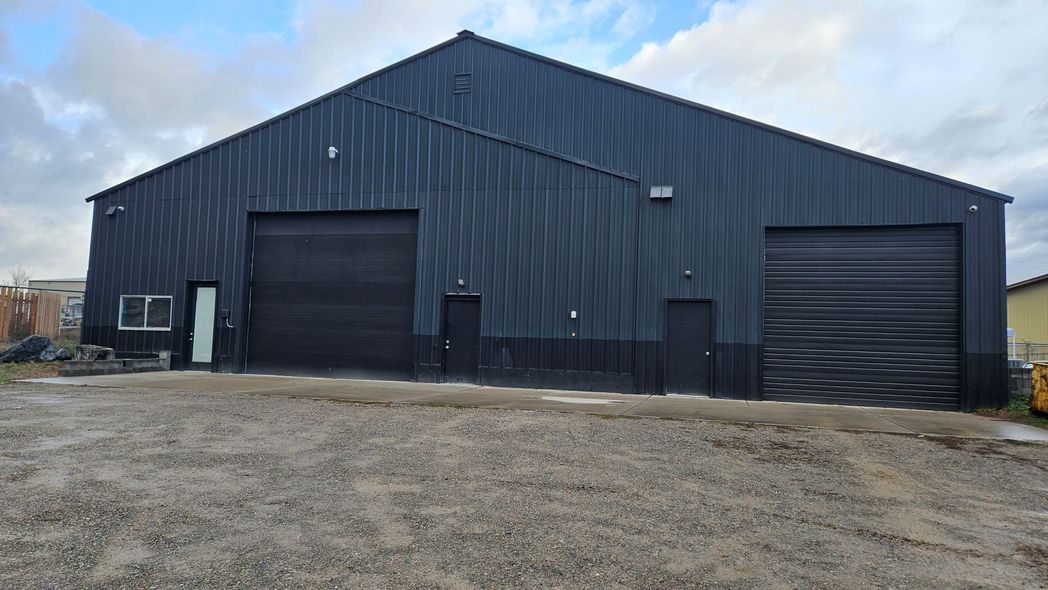 Black industrial building with two large garage doors, two smaller doors, and a window; gravel lot.