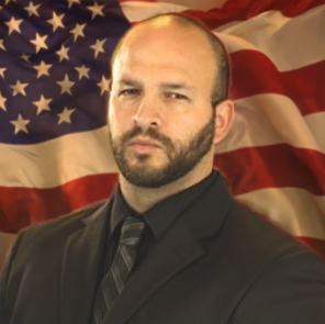 Man in black suit before a red flag backdrop, standing with a serious expression