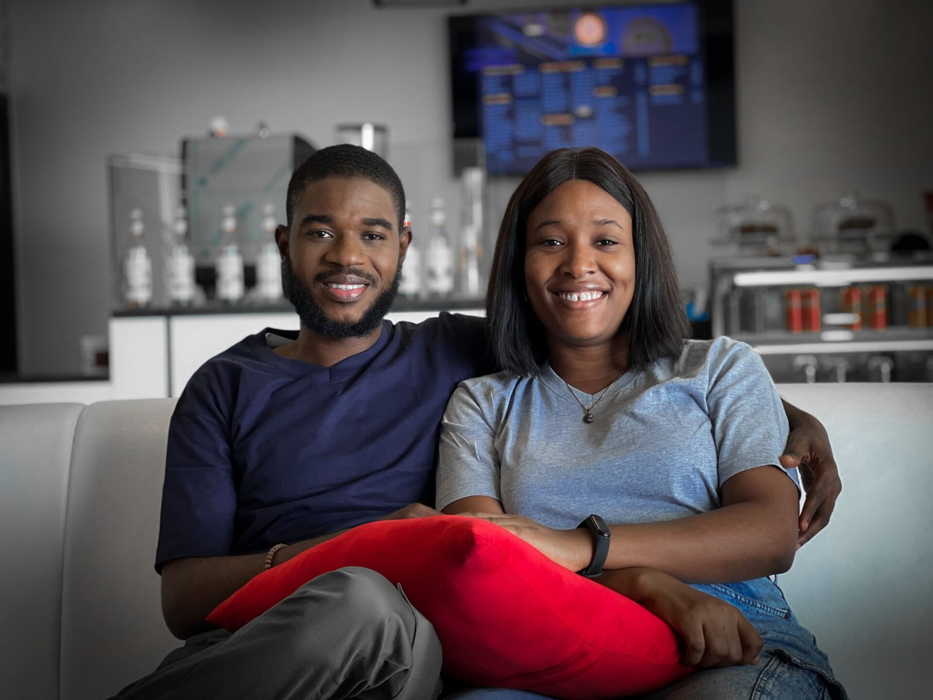 A smiling couple sitting on a white sofa with a red pillow, in front of a counter and a screen.