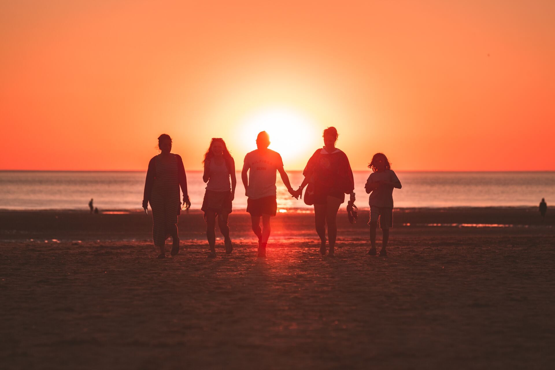 Five people hold hands walking along a sandy beach toward a bright, golden sunset over the ocean.
