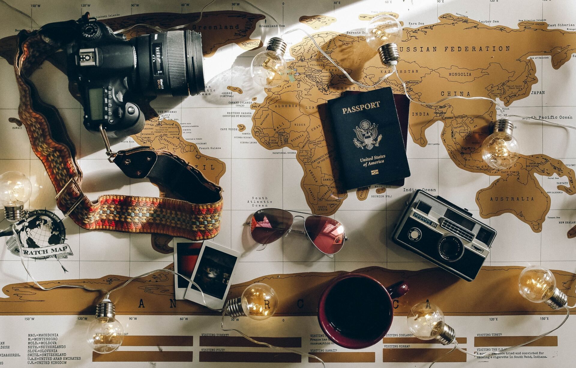 A flat lay of a world map featuring a camera, a US passport, sunglasses, small photos, and fairy lights.