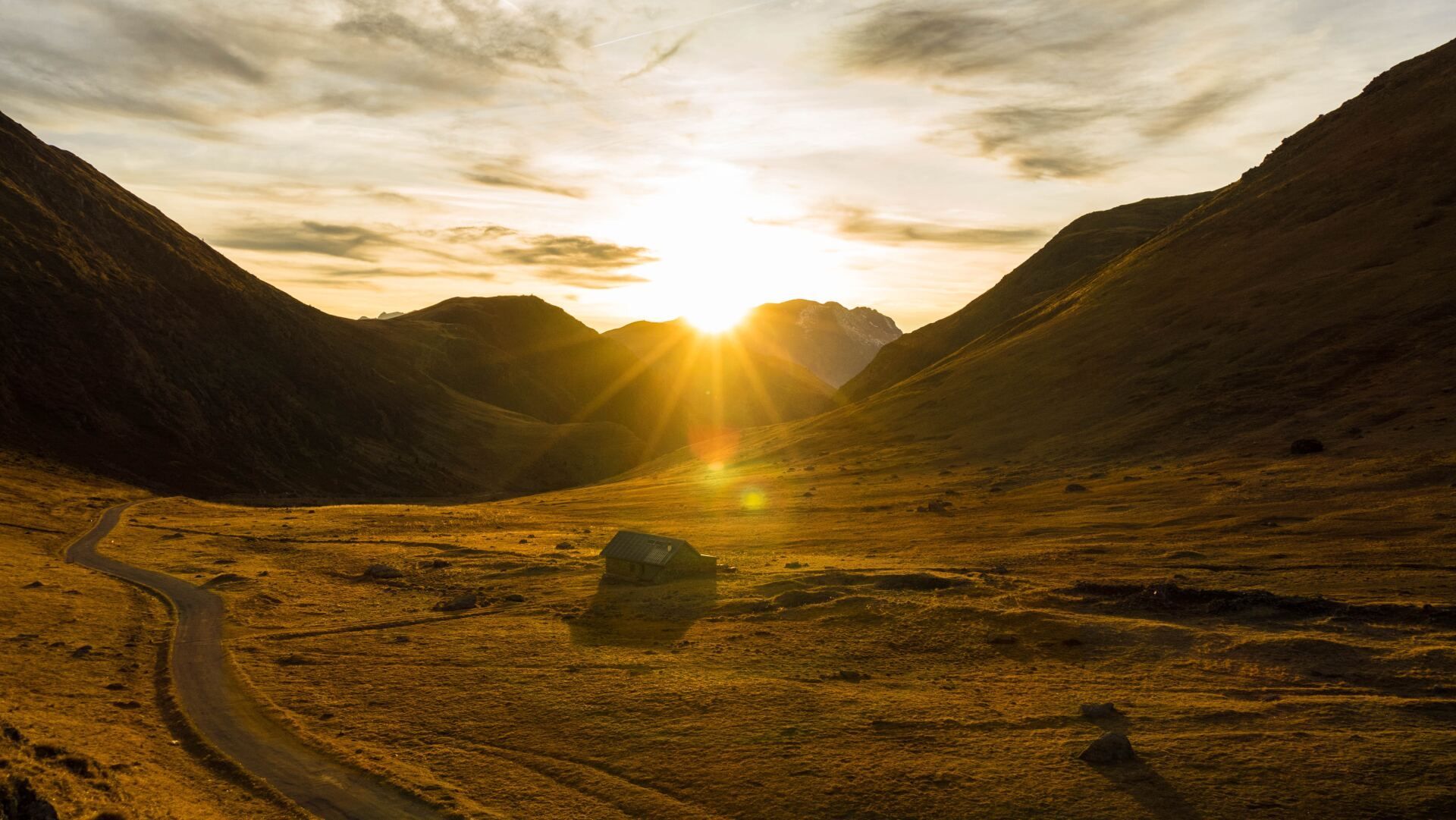 Golden sunset beams over a mountain valley with a winding dirt road and a small building in the foreground.