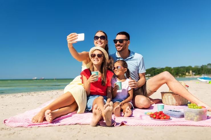 Una familia está sentada sobre una manta en la playa tomándose una selfie.