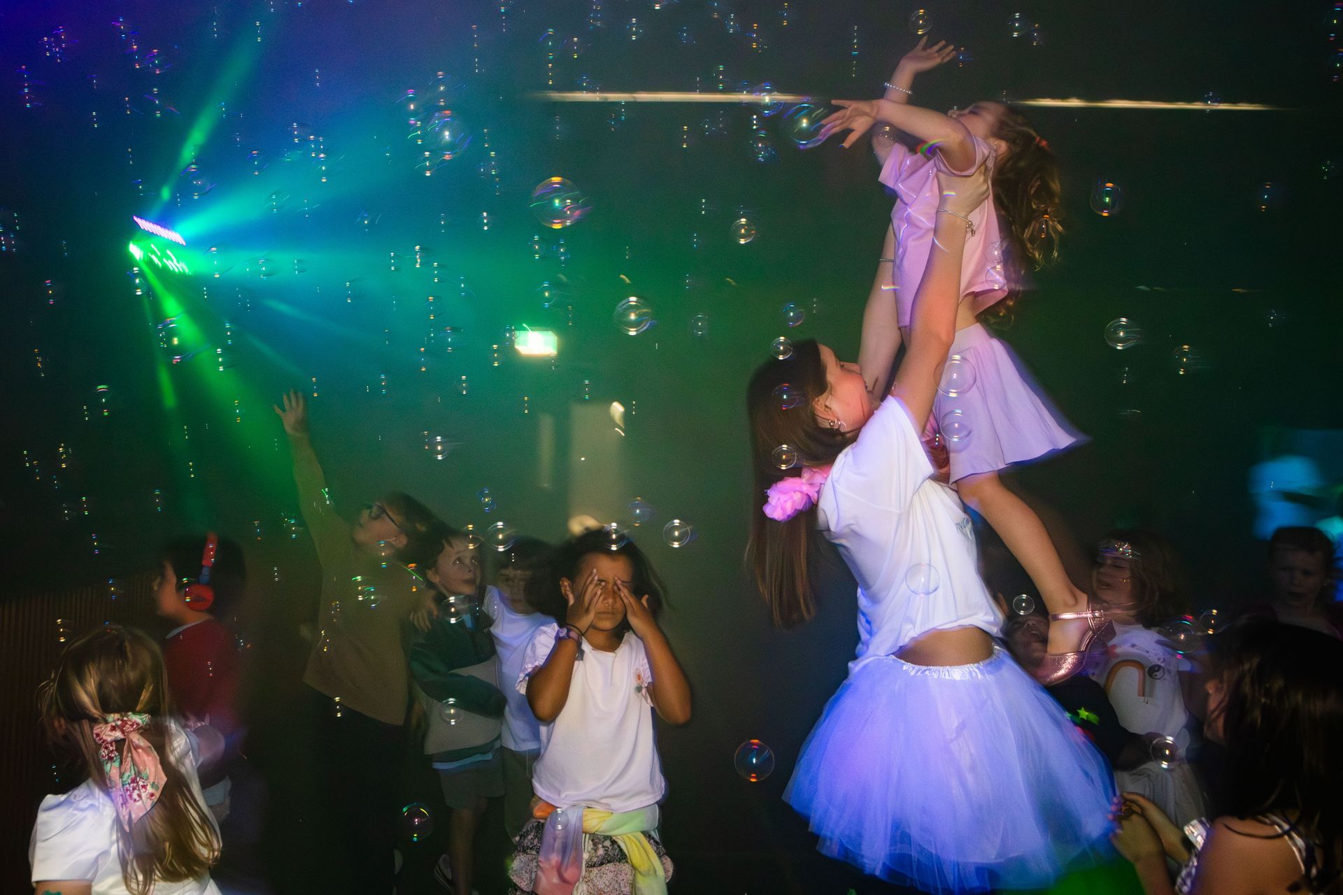 Children playing at a party, one is lifted in the air among bubbles and colorful lights.