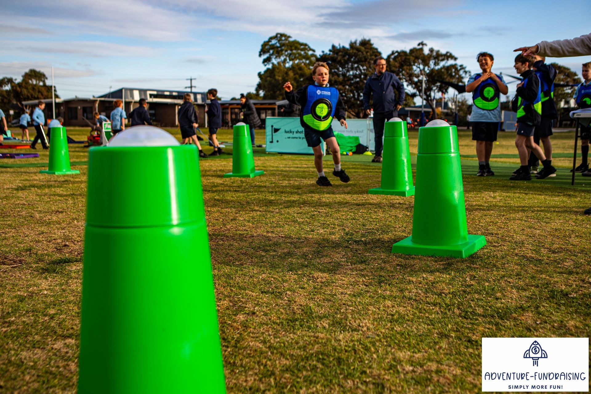 Children playing a game with green cones on a grassy field. People are watching.
